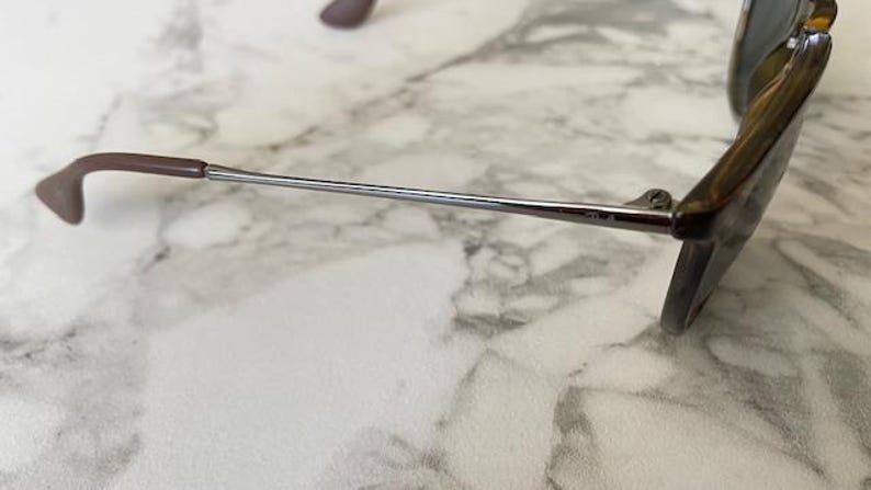 K&ouml;nnte beinhalten: Nahaufnahme einer Sonnenbrille mit braunem Gestell und B&uuml;geln sowie einem silberfarbenen Metallarm. Die Gl&auml;ser sind dunkel gef&auml;rbt. Die Sonnenbrille liegt auf einer Marmoroberfl&auml;che.