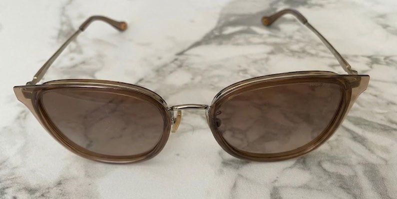 May include: Cat-eye sunglasses with a light brown frame and gradient lenses. The frames are a translucent brown with a silver bridge and brown temples. The lenses transition from a darker brown at the top to a lighter shade at the bottom.