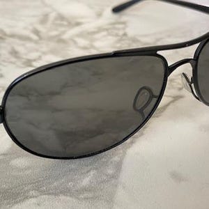 May include: Black aviator sunglasses with dark tinted lenses. The sunglasses have a sleek, modern design with a thin metal frame and a double bridge. The lenses are a dark gray color, providing shade and style.
