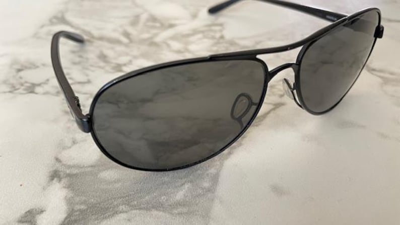 May include: Black aviator sunglasses with dark tinted lenses. The sunglasses have a sleek, modern design with a thin metal frame and a double bridge. The lenses are a dark gray color, providing shade and style.