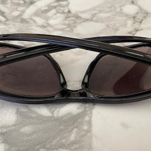 May include: Black sunglasses with dark lenses are folded and resting on a marble surface. The sunglasses have a sleek, modern design with a glossy finish. The lenses appear to be tinted, providing shade and protection from the sun.