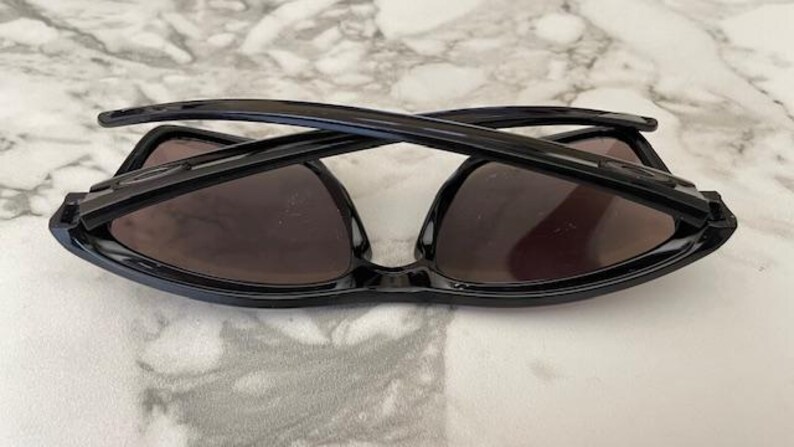 May include: Black sunglasses with dark lenses are folded and resting on a marble surface. The sunglasses have a sleek, modern design with a glossy finish. The lenses appear to be tinted, providing shade and protection from the sun.