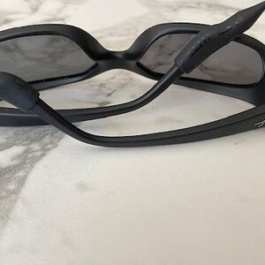 May include: Matte black sunglasses with dark tinted lenses and a black rubberized strap attached to the temples. The Ray-Ban logo is visible in white lettering on the outer side of each temple arm, featuring a wrap-around frame design.