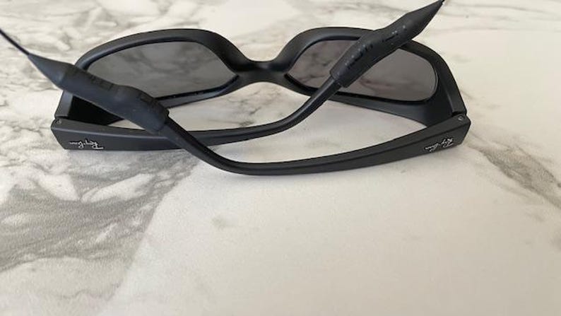May include: Matte black sunglasses with dark tinted lenses and a black rubberized strap attached to the temples. The Ray-Ban logo is visible in white lettering on the outer side of each temple arm, featuring a wrap-around frame design.