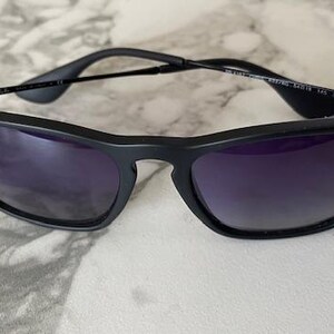 May include: Black sunglasses with dark purple lenses. The sunglasses have a sleek, modern design with a matte finish. The Ray-Ban logo is visible on the left lens. The frame is thin and the arms are black.
