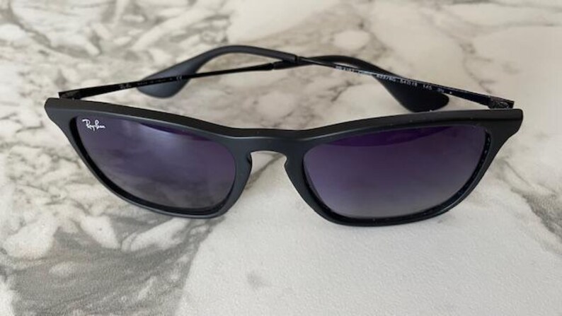 May include: Black sunglasses with dark purple lenses. The sunglasses have a sleek, modern design with a matte finish. The Ray-Ban logo is visible on the left lens. The frame is thin and the arms are black.