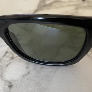 Puede incluir: Primer plano de gafas de sol negras con lentes oscuras. Las gafas de sol tienen una montura gruesa y redondeada y un dise&ntilde;o elegante y moderno. Las lentes son de color verde oscuro y la montura es brillante.