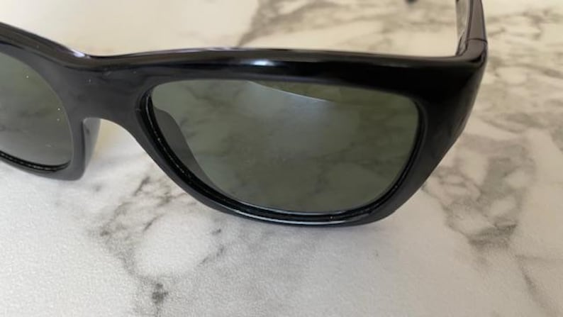 Puede incluir: Primer plano de gafas de sol negras con lentes oscuras. Las gafas de sol tienen una montura gruesa y redondeada y un dise&ntilde;o elegante y moderno. Las lentes son de color verde oscuro y la montura es brillante.