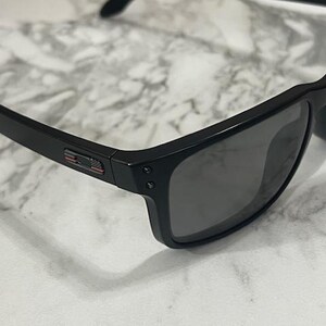 May include: Black sunglasses with dark lenses. The frames are a matte black with a small red, white, and blue accent on the temple. The sunglasses have a classic, rectangular shape.