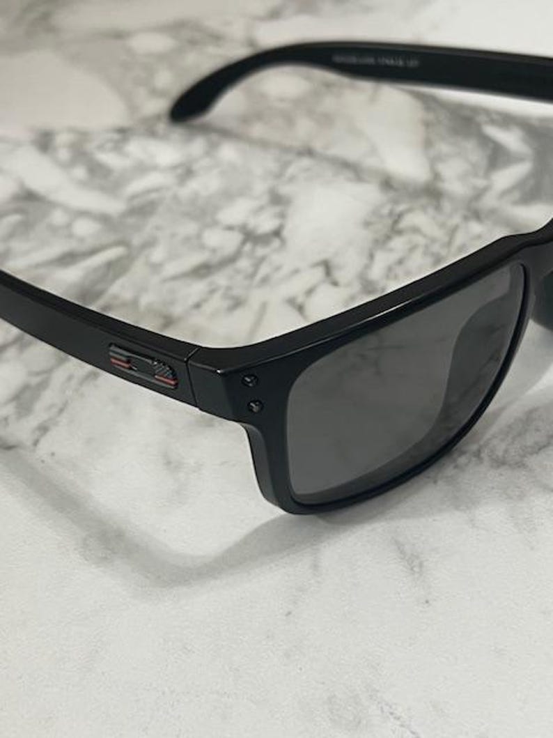 May include: Black sunglasses with dark lenses. The frames are a matte black with a small red, white, and blue accent on the temple. The sunglasses have a classic, rectangular shape.
