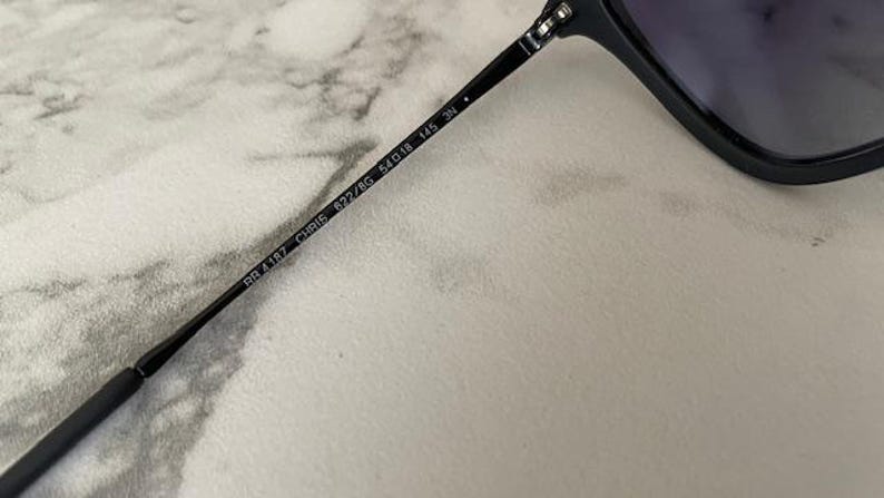 May include: Close-up of a black sunglasses arm with the text "RB 4187 CHRIS 622/8G 54o18 145 3N". The sunglasses arm is black and has a sleek, modern design. The lens is visible in the background.