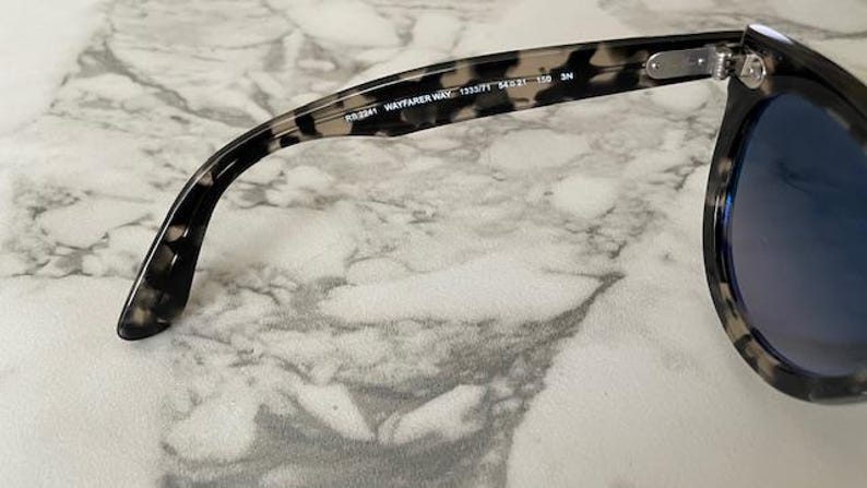 Puede incluir: Primer plano de gafas de sol Wayfarer con un patr&oacute;n de carey en la montura. Las lentes son de color azul oscuro. El texto "RB2141 WAYFARER WAY 1333/71 54 21 150 3N" est&aacute; impreso en la patilla.