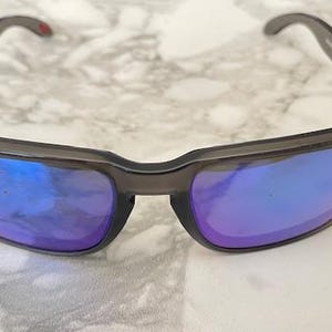 May include: Dark gray sunglasses with blue and purple reflective lenses. The sunglasses have a rectangular frame and a small red accent on the temple. The lenses reflect the surrounding light.