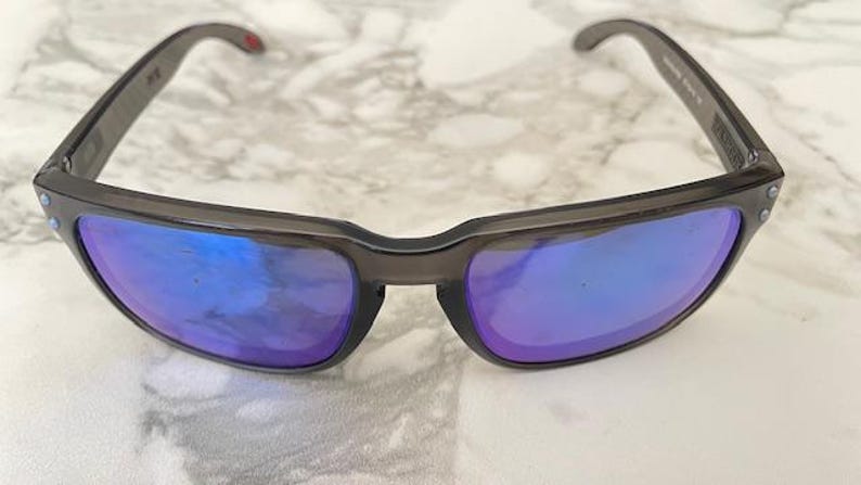 May include: Dark gray sunglasses with blue and purple reflective lenses. The sunglasses have a rectangular frame and a small red accent on the temple. The lenses reflect the surrounding light.