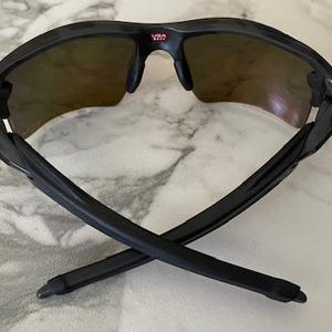 Peut inclure: Lunettes de soleil de sport noires avec une monture incurv&eacute;e et des verres teint&eacute;s. Le pont porte le mot "USA" en rouge. Les branches des lunettes sont crois&eacute;es, mettant en valeur leur design &eacute;pur&eacute;. Ces lunettes sont con&ccedil;ues pour les activit&eacute;s de plein air.