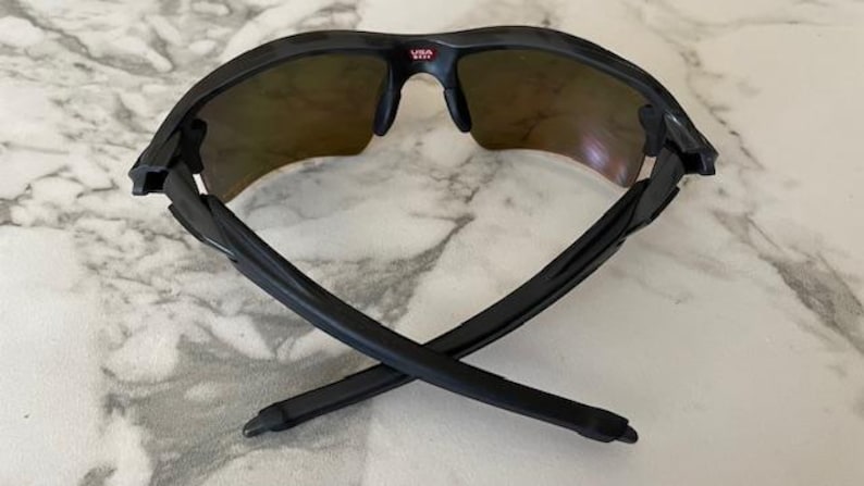 Peut inclure: Lunettes de soleil de sport noires avec une monture incurv&eacute;e et des verres teint&eacute;s. Le pont porte le mot "USA" en rouge. Les branches des lunettes sont crois&eacute;es, mettant en valeur leur design &eacute;pur&eacute;. Ces lunettes sont con&ccedil;ues pour les activit&eacute;s de plein air.