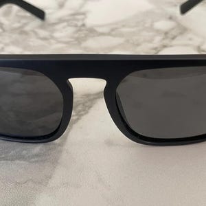 Puede incluir: Gafas de sol en negro mate con una barra superior gruesa y plana y lentes oscuras tintadas. La montura presenta un dise&ntilde;o angular y estructurado con un puente que conecta ambos cristales, ofreciendo un aspecto audaz de l&iacute;neas definidas.