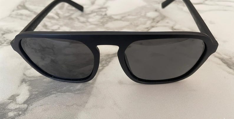 Puede incluir: Gafas de sol en negro mate con una barra superior gruesa y plana y lentes oscuras tintadas. La montura presenta un dise&ntilde;o angular y estructurado con un puente que conecta ambos cristales, ofreciendo un aspecto audaz de l&iacute;neas definidas.