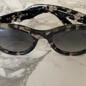 Puede incluir: Gafas de sol estilo Wayfarer con estampado de carey y lentes oscuras. La montura es moteada en negro y marr&oacute;n. El logotipo de Ray-Ban es visible en la lente. Las gafas de sol est&aacute;n sobre una superficie de m&aacute;rmol.