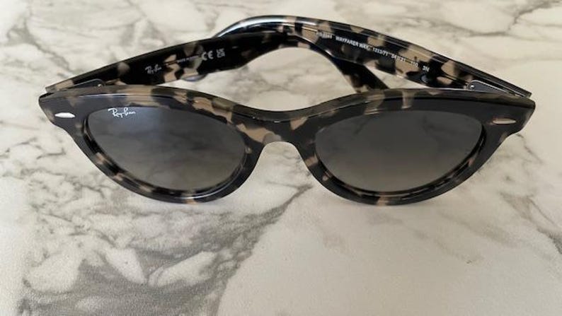 Puede incluir: Gafas de sol estilo Wayfarer con estampado de carey y lentes oscuras. La montura es moteada en negro y marr&oacute;n. El logotipo de Ray-Ban es visible en la lente. Las gafas de sol est&aacute;n sobre una superficie de m&aacute;rmol.