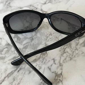 K&ouml;nnte beinhalten: Schwarze Sonnenbrille mit dunklen Gl&auml;sern. Die Sonnenbrille hat einen dicken, abgerundeten Rahmen, und das Ray-Ban-Logo ist auf dem B&uuml;gel sichtbar. Die B&uuml;gel sind schlank und erstrecken sich nach hinten, was einen klassischen, stilvollen Look erzeugt.