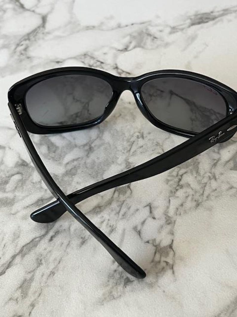 K&ouml;nnte beinhalten: Schwarze Sonnenbrille mit dunklen Gl&auml;sern. Die Sonnenbrille hat einen dicken, abgerundeten Rahmen, und das Ray-Ban-Logo ist auf dem B&uuml;gel sichtbar. Die B&uuml;gel sind schlank und erstrecken sich nach hinten, was einen klassischen, stilvollen Look erzeugt.