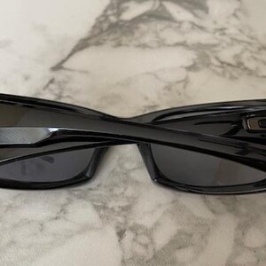K&ouml;nnte beinhalten: Schwarze Sonnenbrille mit gl&auml;nzendem Finish, mit rechteckigem Rahmen und dunklen Gl&auml;sern. Die B&uuml;gel haben ein markantes Design mit metallischem Akzent. Die Sonnenbrille liegt auf einer Marmoroberfl&auml;che.