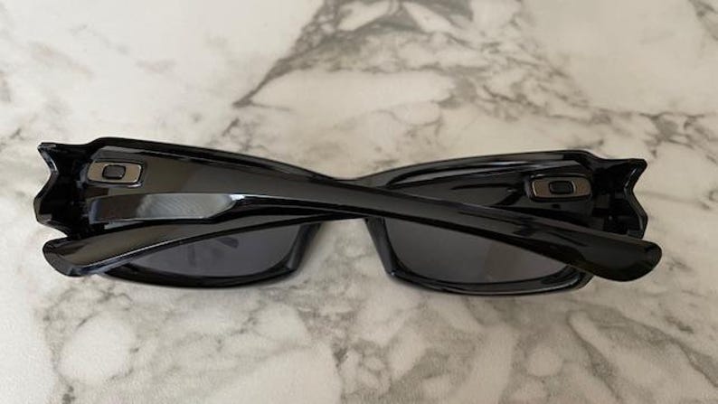 K&ouml;nnte beinhalten: Schwarze Sonnenbrille mit gl&auml;nzendem Finish, mit rechteckigem Rahmen und dunklen Gl&auml;sern. Die B&uuml;gel haben ein markantes Design mit metallischem Akzent. Die Sonnenbrille liegt auf einer Marmoroberfl&auml;che.