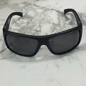 May include: Black sunglasses with a wraparound design. The sunglasses have dark lenses and a thick, matte black frame. The temples curve slightly for a comfortable fit. The sunglasses are set against a white and gray marble background.