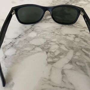 May include: Black sunglasses with dark lenses. The sunglasses have a classic rectangular frame and are set against a marble background. The arms of the sunglasses are extended, showcasing the design.