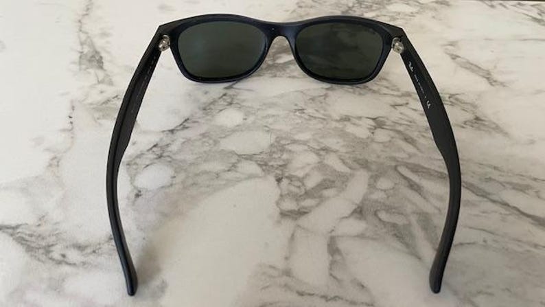 May include: Black sunglasses with dark lenses. The sunglasses have a classic rectangular frame and are set against a marble background. The arms of the sunglasses are extended, showcasing the design.