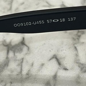 May include: Close-up of the inner arm of black sunglasses. The text "OO9102-U455 57 18 137" is printed in white on the black frame. The background is a light gray and white marbled surface.