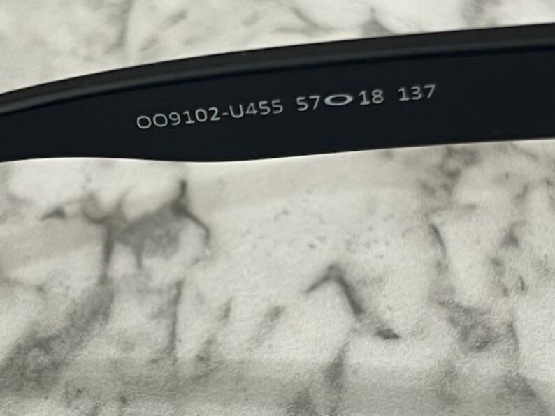 May include: Close-up of the inner arm of black sunglasses. The text "OO9102-U455 57 18 137" is printed in white on the black frame. The background is a light gray and white marbled surface.