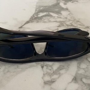 Puede incluir: Gafas de sol negras con lentes azul oscuro plegadas sobre una superficie de m&aacute;rmol. Las gafas de sol tienen un acabado brillante y el logotipo de Ray-Ban es visible en las patillas. El dise&ntilde;o es elegante y moderno.