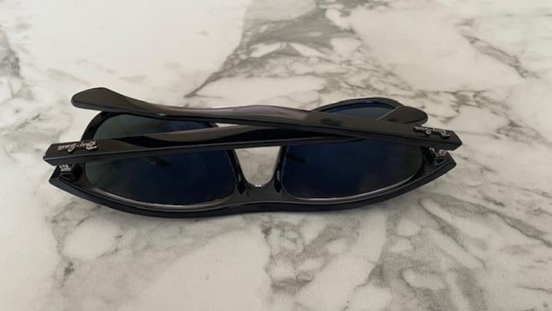 Puede incluir: Gafas de sol negras con lentes azul oscuro plegadas sobre una superficie de m&aacute;rmol. Las gafas de sol tienen un acabado brillante y el logotipo de Ray-Ban es visible en las patillas. El dise&ntilde;o es elegante y moderno.