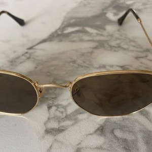 May include: Gold-framed sunglasses with brown lenses. The Ray-Ban logo is visible on the left lens. The glasses have a classic, elegant design with thin temples and a bridge connecting the lenses. The sunglasses are set against a marble background.