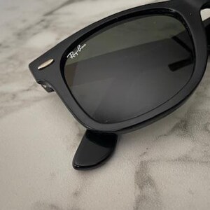 K&ouml;nnte beinhalten: Schwarze Sonnenbrille mit dunklen Gl&auml;sern. Die Sonnenbrille hat ein klassisches rechteckiges Rahmendesign. Das Ray-Ban-Logo ist auf dem oberen linken Glas sichtbar. Die Sonnenbrille ist vor einem hellen, marmorierten Hintergrund platziert.