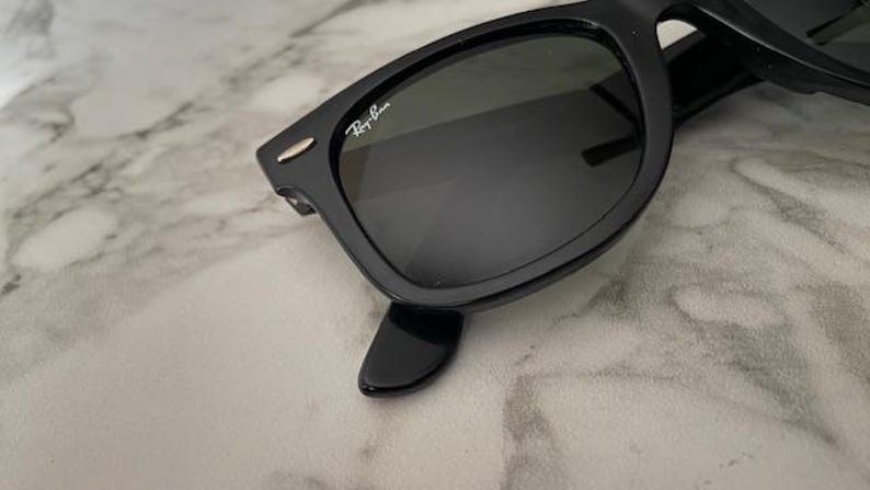 K&ouml;nnte beinhalten: Schwarze Sonnenbrille mit dunklen Gl&auml;sern. Die Sonnenbrille hat ein klassisches rechteckiges Rahmendesign. Das Ray-Ban-Logo ist auf dem oberen linken Glas sichtbar. Die Sonnenbrille ist vor einem hellen, marmorierten Hintergrund platziert.