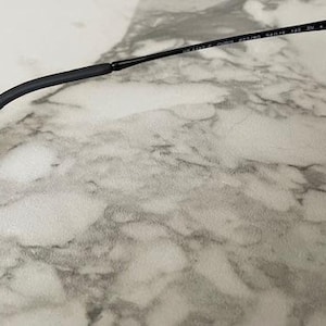 Puede incluir: Gafas de sol negras con lentes oscuras y montura negra. La patilla es curva y tiene un extremo negro. Las gafas de sol est&aacute;n sobre una superficie de m&aacute;rmol.