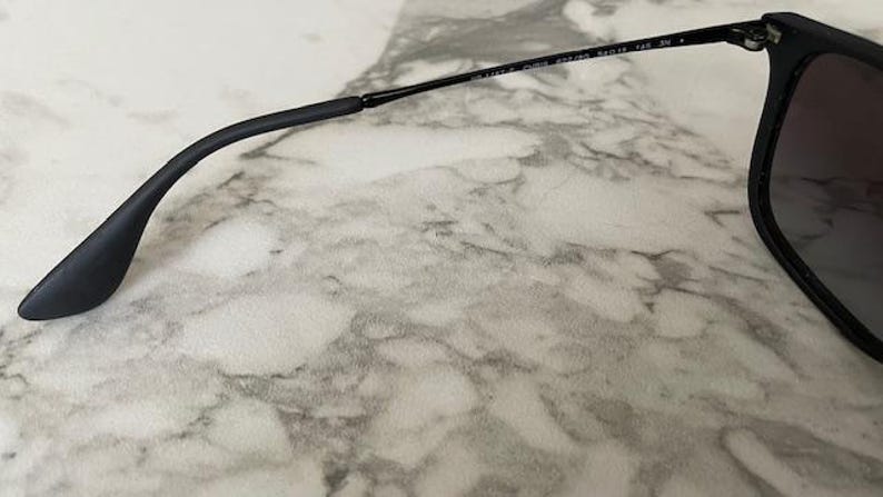 Puede incluir: Gafas de sol negras con lentes oscuras y montura negra. La patilla es curva y tiene un extremo negro. Las gafas de sol est&aacute;n sobre una superficie de m&aacute;rmol.