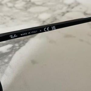 Puede incluir: Primer plano de gafas de sol negras con el logotipo de Ray-Ban y el texto "MADE IN ITALY". Las gafas de sol tienen un dise&ntilde;o elegante y est&aacute;n marcadas con "CE" y "UK CA".