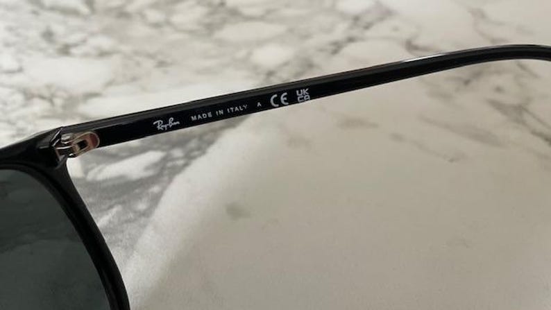 Puede incluir: Primer plano de gafas de sol negras con el logotipo de Ray-Ban y el texto "MADE IN ITALY". Las gafas de sol tienen un dise&ntilde;o elegante y est&aacute;n marcadas con "CE" y "UK CA".
