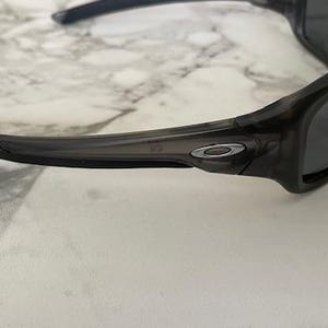 May include: Dark gray sunglasses with a wraparound design. The sunglasses have a black frame and dark lenses. The Oakley logo is visible on the temples. The sunglasses are designed for sports and outdoor activities.