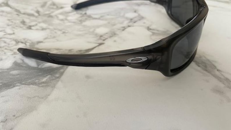 May include: Dark gray sunglasses with a wraparound design. The sunglasses have a black frame and dark lenses. The Oakley logo is visible on the temples. The sunglasses are designed for sports and outdoor activities.