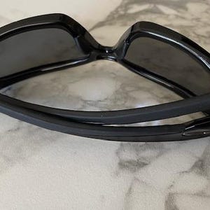 May include: Black sunglasses with a glossy finish. The sunglasses have a rectangular frame and dark lenses. The arms are curved and have a rubberized grip. The sunglasses are designed for outdoor use and protection from the sun.