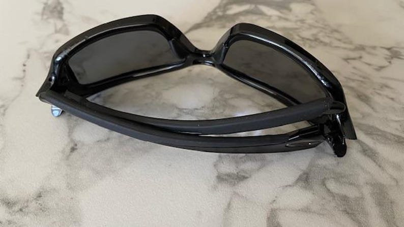 May include: Black sunglasses with a glossy finish. The sunglasses have a rectangular frame and dark lenses. The arms are curved and have a rubberized grip. The sunglasses are designed for outdoor use and protection from the sun.