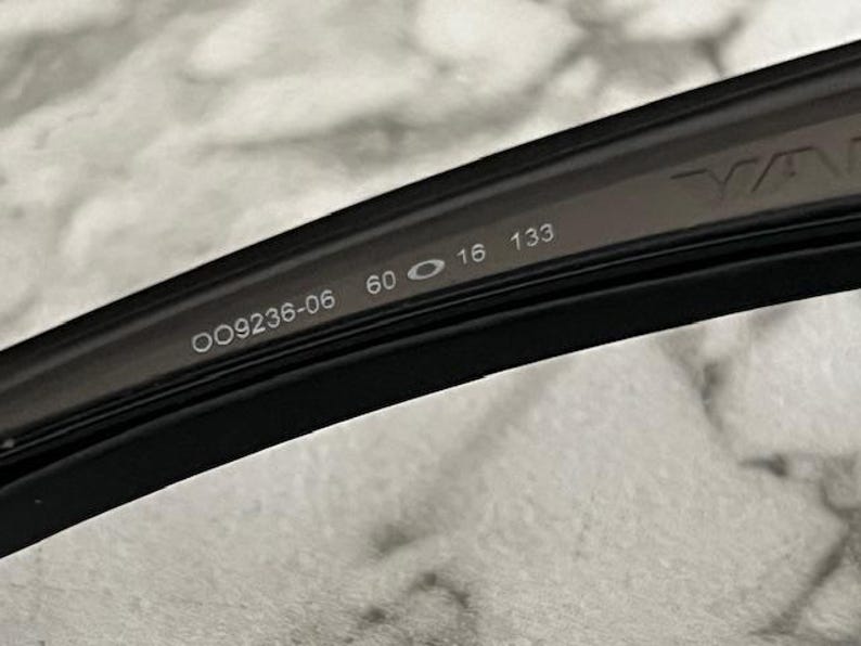 May include: Close-up of a black eyeglass frame with the model number OO9236-06, lens width 60 mm, bridge width 16 mm, and temple length 133 mm. The brand name is partially visible.