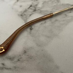 May include: Close-up of a brown and gold eyeglass arm. The arm is curved and has a gold "C" logo at the end. The arm is connected to a thin, silver metal frame. The background is a light gray and white marble pattern.