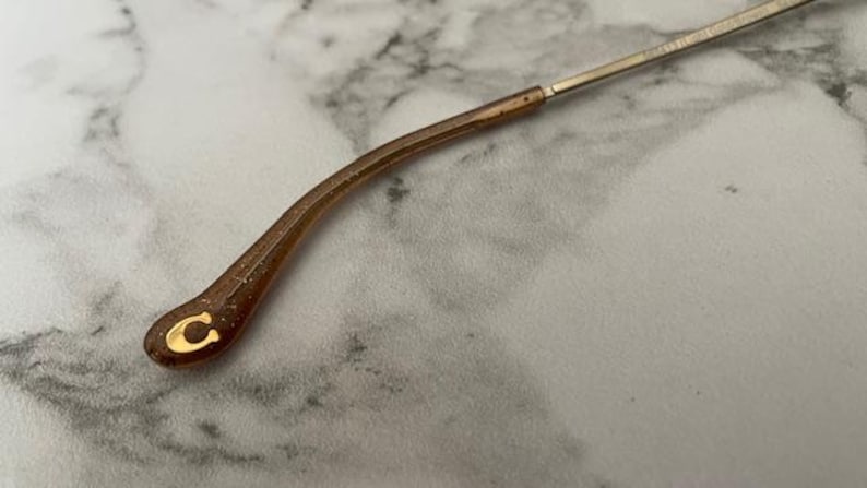 May include: Close-up of a brown and gold eyeglass arm. The arm is curved and has a gold "C" logo at the end. The arm is connected to a thin, silver metal frame. The background is a light gray and white marble pattern.