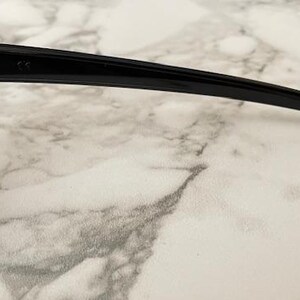 May include: Close-up of black sunglasses with a rectangular frame and dark lenses. The sunglasses have a sleek, modern design with a curved arm. The image is set against a marble-like background.